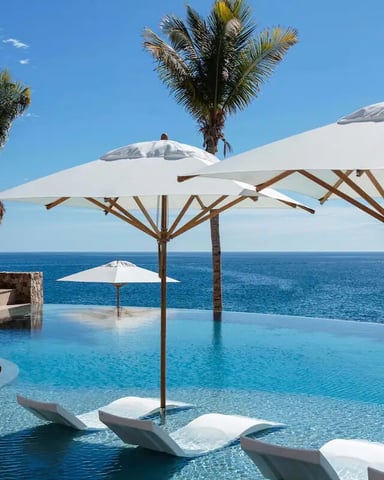Infinity pool with white umbrellas overlooking ocean, palm trees, and thatched-roof structures