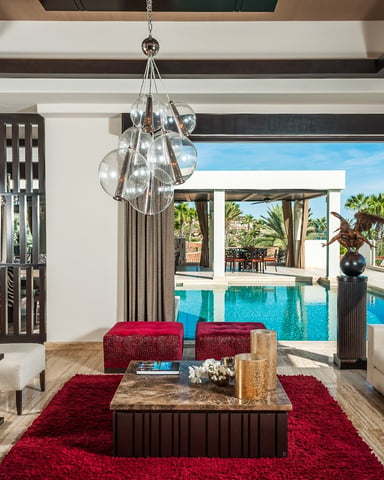 Luxury living room with red accents, modern chandelier, and ocean view through open glass doors to pool and palm trees