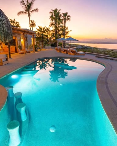 Illuminated curved pool at sunset with modern house, palm trees, and ocean coastline view at dusk