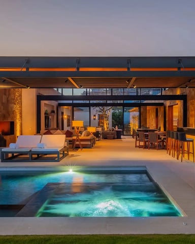 Modern patio at dusk with illuminated pool, lounge chairs, dining area, and open-plan living space with warm lighting