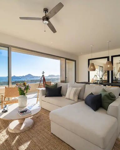 Modern living room with cream sectional sofa, ocean and mountain views through floor-to-ceiling windows, ceiling fan, and pendant lights over kitchen area