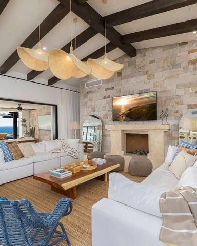 Modern living room with exposed dark beams, stone fireplace, white sofas, blue woven chairs, and ocean view through floor-to-ceiling windows