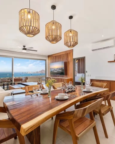Open-plan living and dining area with wooden table, ocean view through floor-to-ceiling windows, and three pendant lights overhead.