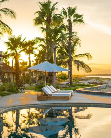 Beachfront resort at sunset with curved pool, lounge chairs, blue umbrella, palm trees, and ocean view