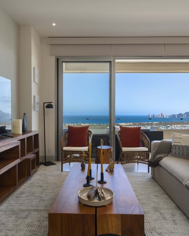 Modern living room with ocean and mountain views through floor-to-ceiling windows, wood furniture, and coastal seating area