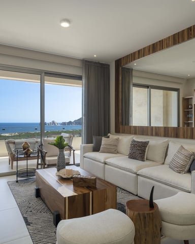 Modern living room with cream sectional sofa, ocean and rocky coastline view through floor-to-ceiling windows, wood accents, and coastal landscape beyond