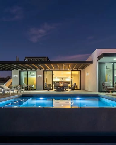 Modern house with illuminated swimming pool, lounge chairs, pergola, and lit interior spaces at dusk