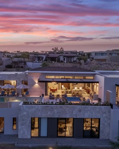 Modern white villa with rooftop pool and terrace at dusk, illuminated interior, Mediterranean hillside setting with distant mountains
