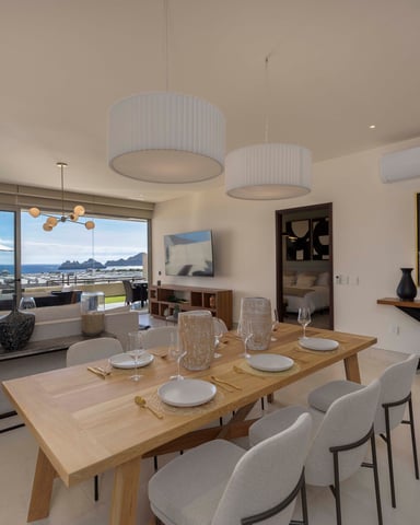 Modern open-plan dining and living room with ocean and mountain views, wooden dining table, pendant lights, and coastal furnishings.
