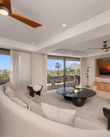 Modern living room with curved white sofa, black round table, wall-mounted TV, ceiling fans, and floor-to-ceiling windows overlooking palm trees and city