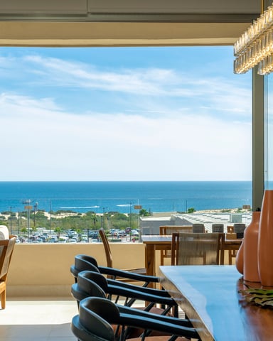 Modern dining room with floor-to-ceiling windows overlooking blue ocean, coastal landscape, and distant rock formations