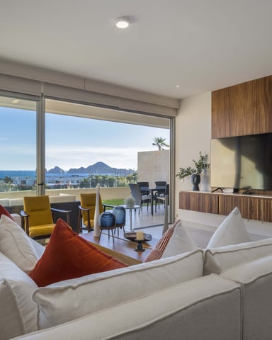Modern living room with cream sectional sofa, wall-mounted TV, and floor-to-ceiling windows overlooking coastal mountains and ocean.