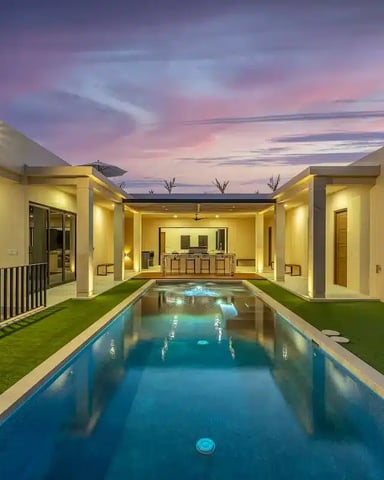Modern courtyard home with illuminated cream-colored walls, rectangular lap pool, artificial turf, and twilight sky at dusk