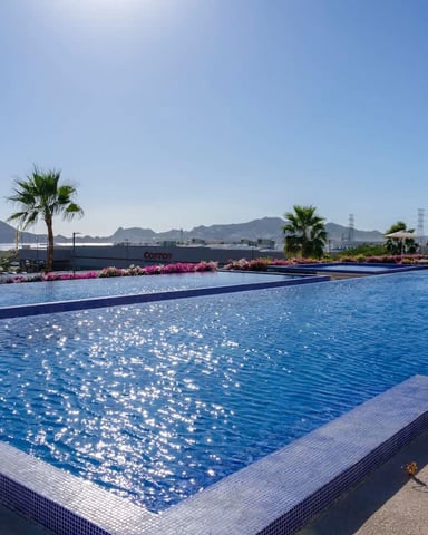 Large resort swimming pool with blue water, palm trees, pink flowers, and desert mountains in background under clear sky
