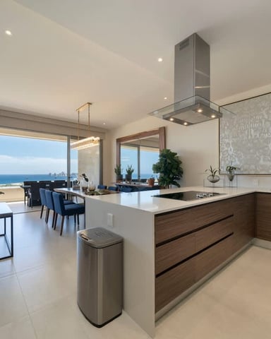 Modern open-plan kitchen and living room with ocean view, white countertops, wood cabinetry, and floor-to-ceiling windows overlooking beach.