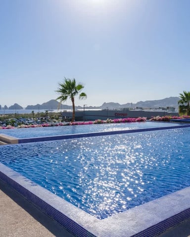 Resort pool with blue water overlooking palm trees, pink flowers, mountains, and coastal bay