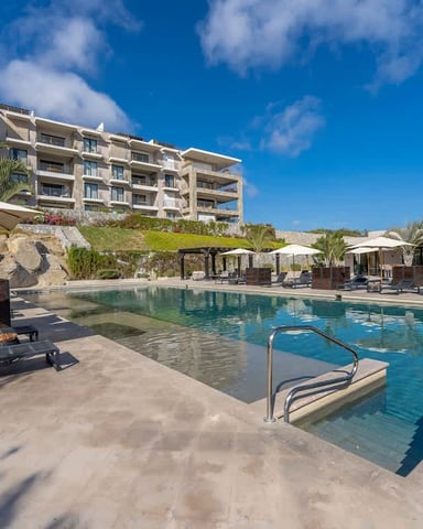 Resort pool with loungers and umbrellas, modern apartment building, palm trees, and clear blue sky