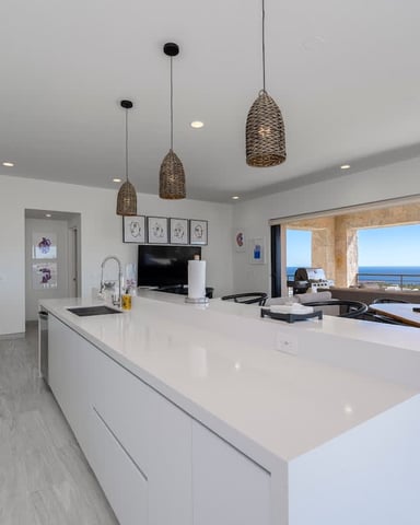Modern open-plan kitchen and living room with white cabinetry, woven pendant lights, and ocean view through large sliding glass doors.