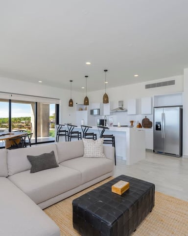 Open-plan living room with beige sectional sofa, kitchen, dining area, and waterfront view through large windows