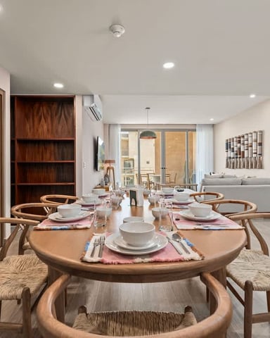 Modern dining room with round wooden table, woven chairs, place settings, and open-plan living area with sofa and balcony beyond.