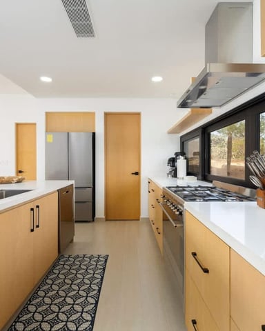 Modern kitchen with natural wood cabinets, white countertops, stainless steel appliances, and large windows overlooking desert landscape.