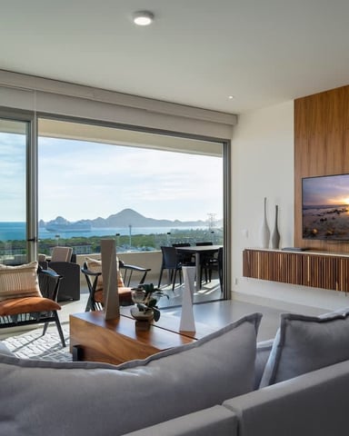 Modern living room with gray seating, wood accent wall, wall-mounted TV, and floor-to-ceiling windows overlooking ocean and distant mountains