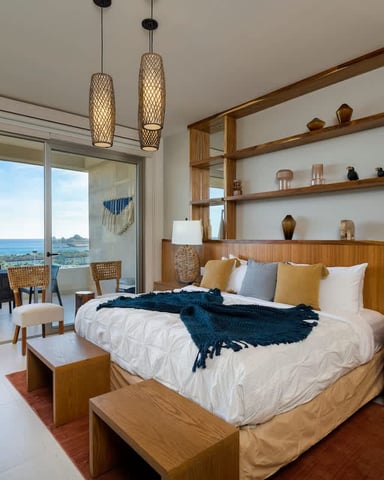 Modern bedroom with wooden furniture, ocean view through sliding glass doors, pendant lights, and built-in shelving.