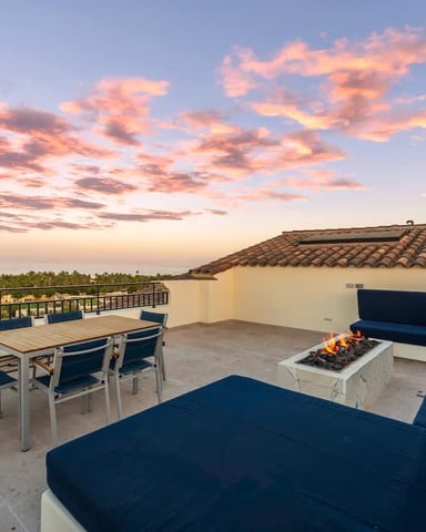 Rooftop terrace at sunset with blue seating, fire pit, dining table, and palm tree views