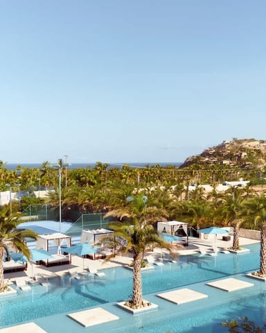 Resort pool complex with turquoise water, palm trees, loungers, and rocky hillside with sea view