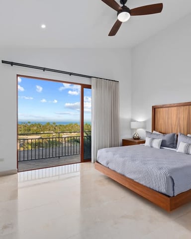 Bedroom with wooden bed, ocean and palm tree view through glass doors, ceiling fan, and coastal landscape visible from balcony