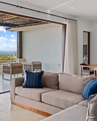 Open-plan living room with beige sofa, ocean and palm tree views through floor-to-ceiling windows, dining area beyond.