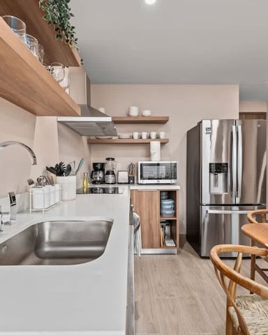Modern kitchen with white countertops, stainless steel sink and appliances, wooden shelving, and dining table with wooden chairs