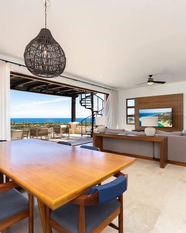 Open-plan living room with wooden dining table, ocean view through large glass doors, and coastal landscape visible from terrace.
