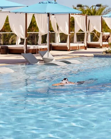Swimmer in resort pool with blue umbrellas, cabanas, and loungers along the deck