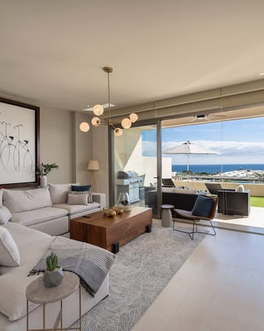 Modern living room with beige sofa, wood coffee table, and floor-to-ceiling windows overlooking ocean and coastal town