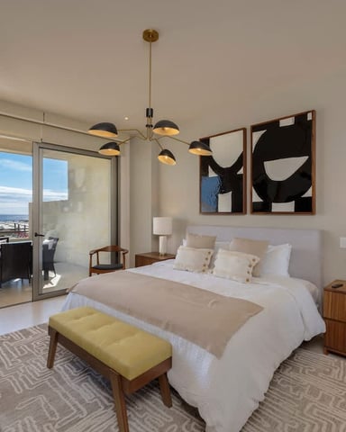 Modern bedroom with white bedding, wood furniture, pendant lighting, and ocean view through glass doors to balcony