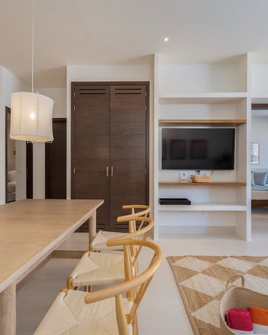 Open-plan living space with light wood dining table, white shelving unit with TV, dark wood closet doors, and pendant lighting