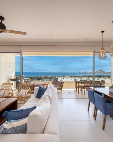 Open-plan living and dining area with ocean and mountain views through floor-to-ceiling windows, white sofa, wood furniture, and pendant lighting.