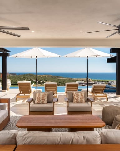 Modern living room with ocean view, opening to terrace with lounge chairs, umbrellas, and swimming pool overlooking blue sea.
