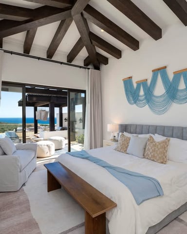 Bright bedroom with exposed wood beams, ocean view through glass doors, blue wall art, and modern furnishings