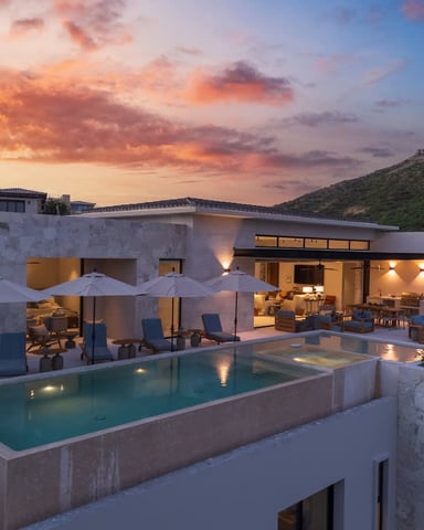 Modern white villa with illuminated pool, lounge chairs, umbrellas, and terrace at sunset with hillside view
