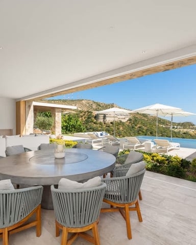 Modern dining area with round concrete table and gray rope chairs, opening to infinity pool and Mediterranean hillside sea view
