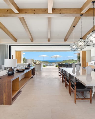 Open-plan kitchen and dining area with exposed wooden beams, white marble countertops, and ocean view through large glass doors.