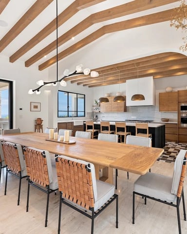 Open-plan dining and kitchen with exposed wood beams, ocean view, and woven leather chairs around wooden table