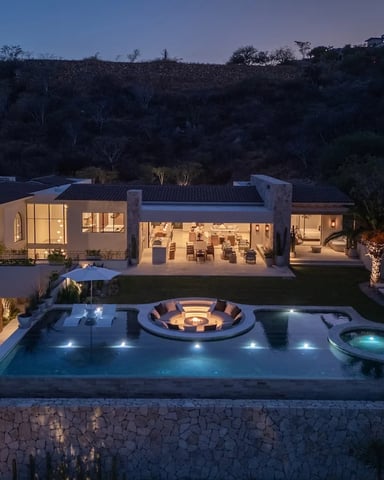 Modern hillside home at dusk with illuminated pool, fire pit, hot tub, and wooded landscape backdrop