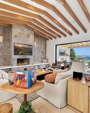 Modern living room with exposed wood beams, stone fireplace, cream seating, and ocean view through open glass doors