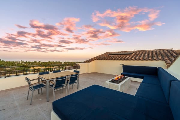 Rooftop terrace at sunset with blue seating, fire pit, dining table, and palm tree views