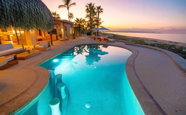 Illuminated curved pool overlooking beach and ocean at sunset, with palm trees, lounge chairs, and modern villa with thatched roof